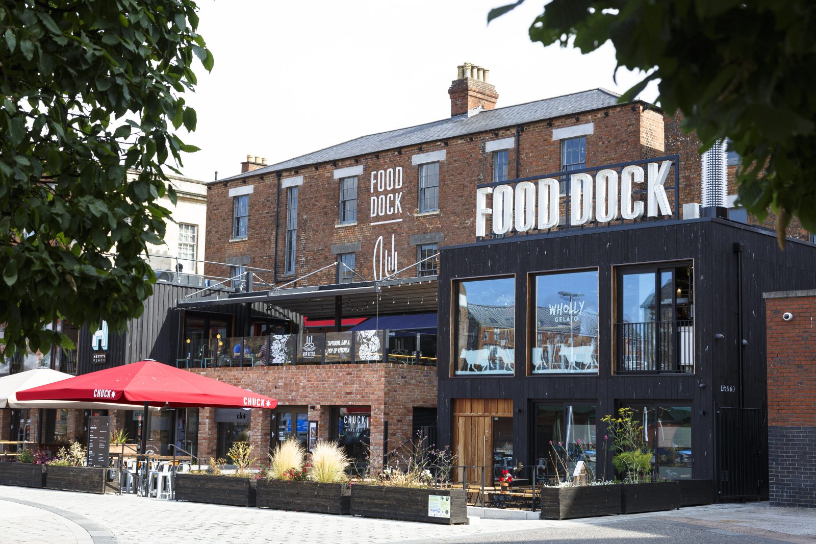 An image through the trees of Gloucester Food Dock. A historic brick building, with modern extensions out the front.